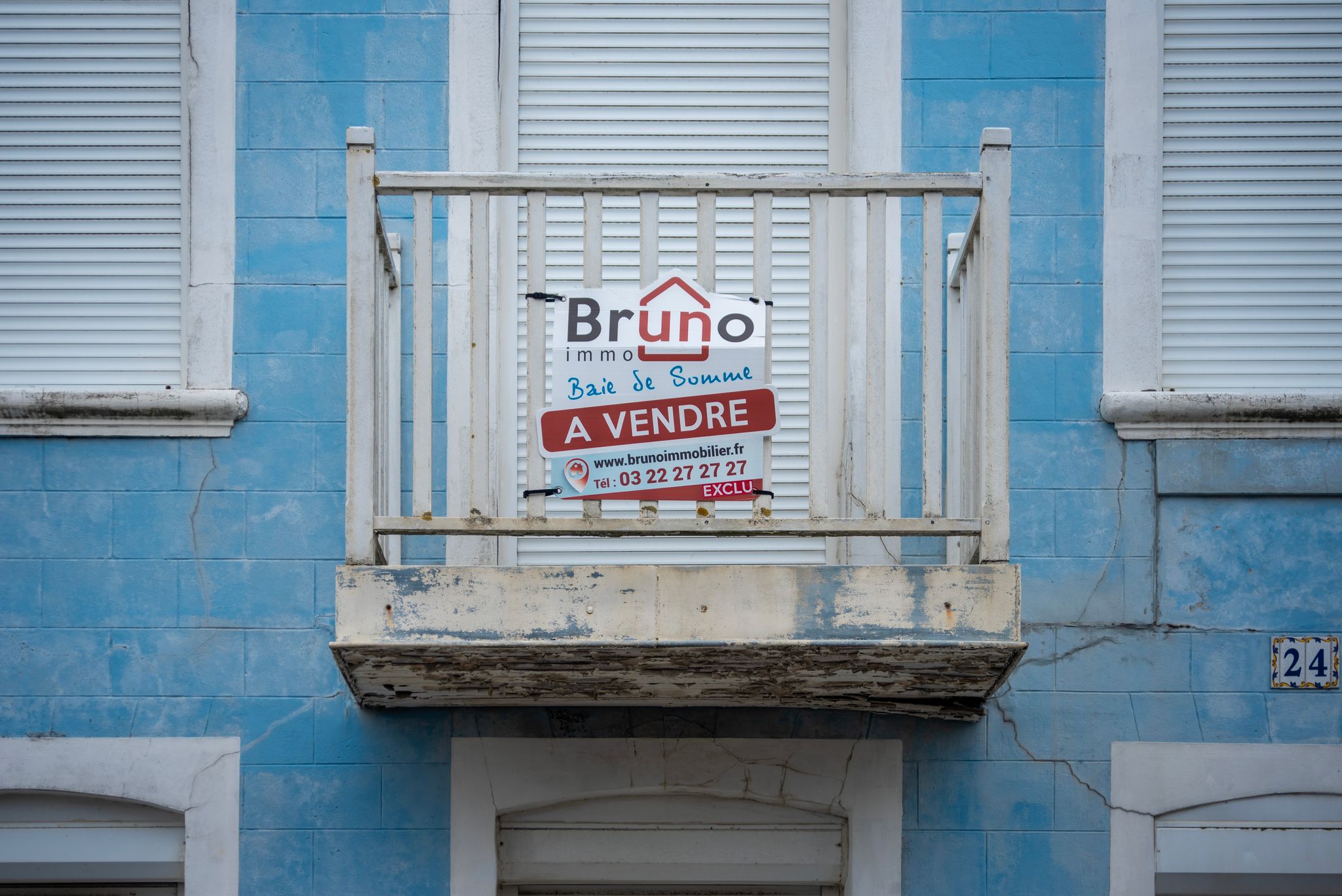 A worn blue facade with a 'For Sale' sign on a rustic balcony.